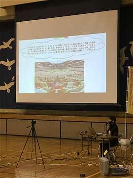 スクリーンの前で講演する横川信子さんの写真
